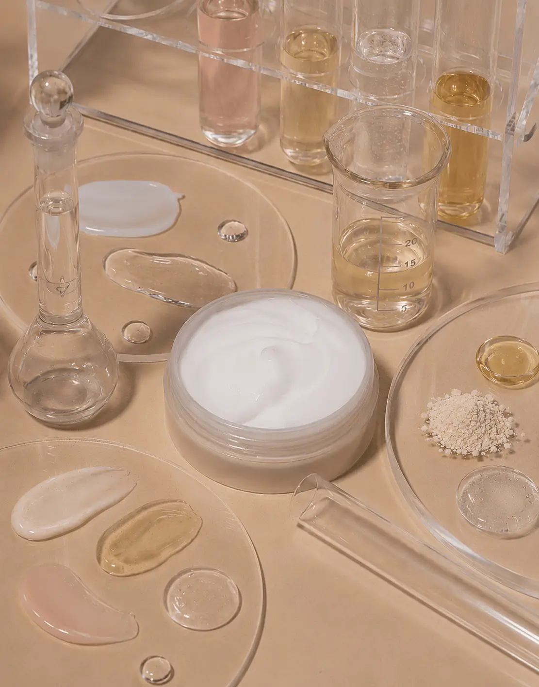 Cream formulation in clear jar surrounded by petri dishes with hyaluronic acid gel, botanical oils, and powdered actives, alongside glass labware and test tubes.