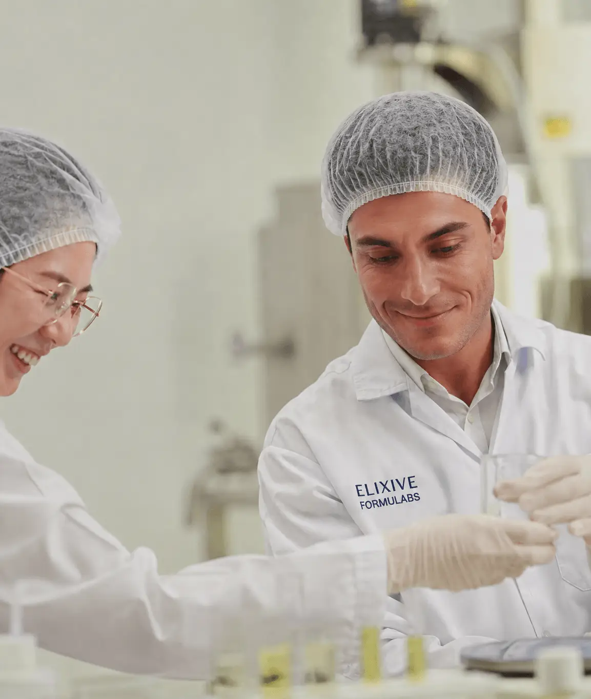 Two Elixive Labs chemists in lab coats and hairnets analyzing clear liquid formulations with glassware, surrounded by vials containing botanical extracts